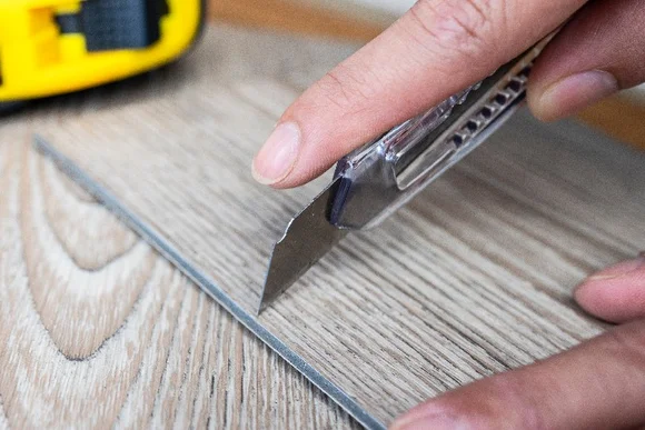 Person will mit Teppichmesser Kleber zwischen Vinylboden entfernen.
